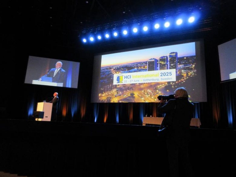 図1、HCI International 2025の会場でスピーカーが登壇している写真。スクリーンにイベント情報と都市の夜景が映り、カメラマンが撮影中。