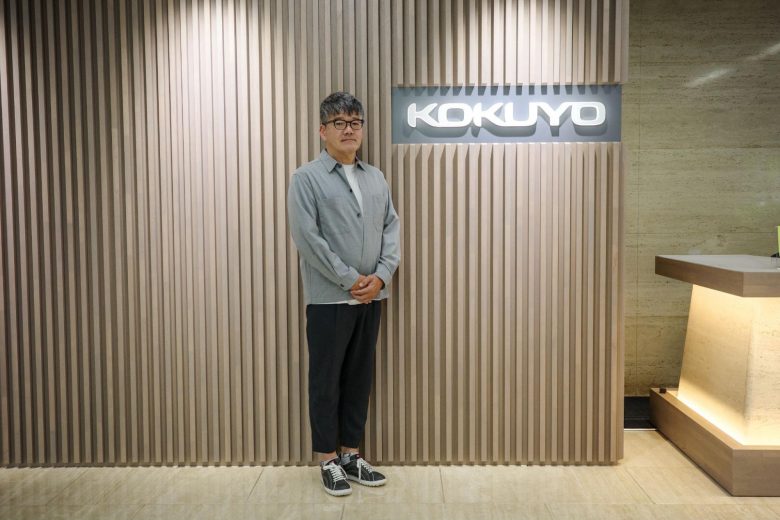 KOKUYOのロゴのある壁の前に人物が立っている。