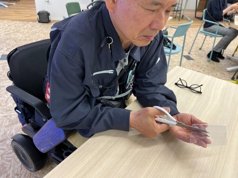 車椅子に座った人物がテーブルの上で透明な物体をハサミで切っている。
