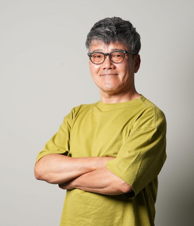 無地の背景の前に、人物が腕を組んで立っている。