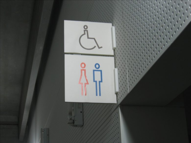 建物内部の入口上部にある案内看板の2つの人物シルエット。左側:上に円形、中央に上向き三角形、下に直線、オレンジの線。右側:上に円形、中央に四角形、下に直線、青の線。