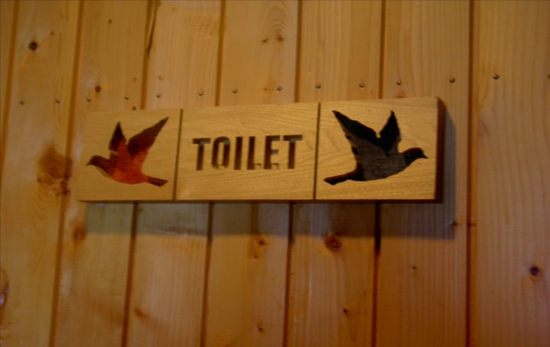 木製の案内板。中央に「TOILET」の文字が彫られ、その左右に飛んでいる鳥のシルエットが彫られている。左の鳥は赤く、右の鳥は黒く塗られている。