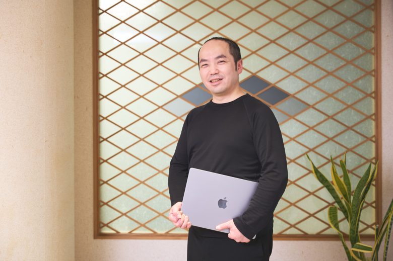 ノートPCを両手でかかえて立っている、笑顔の羽山さん。